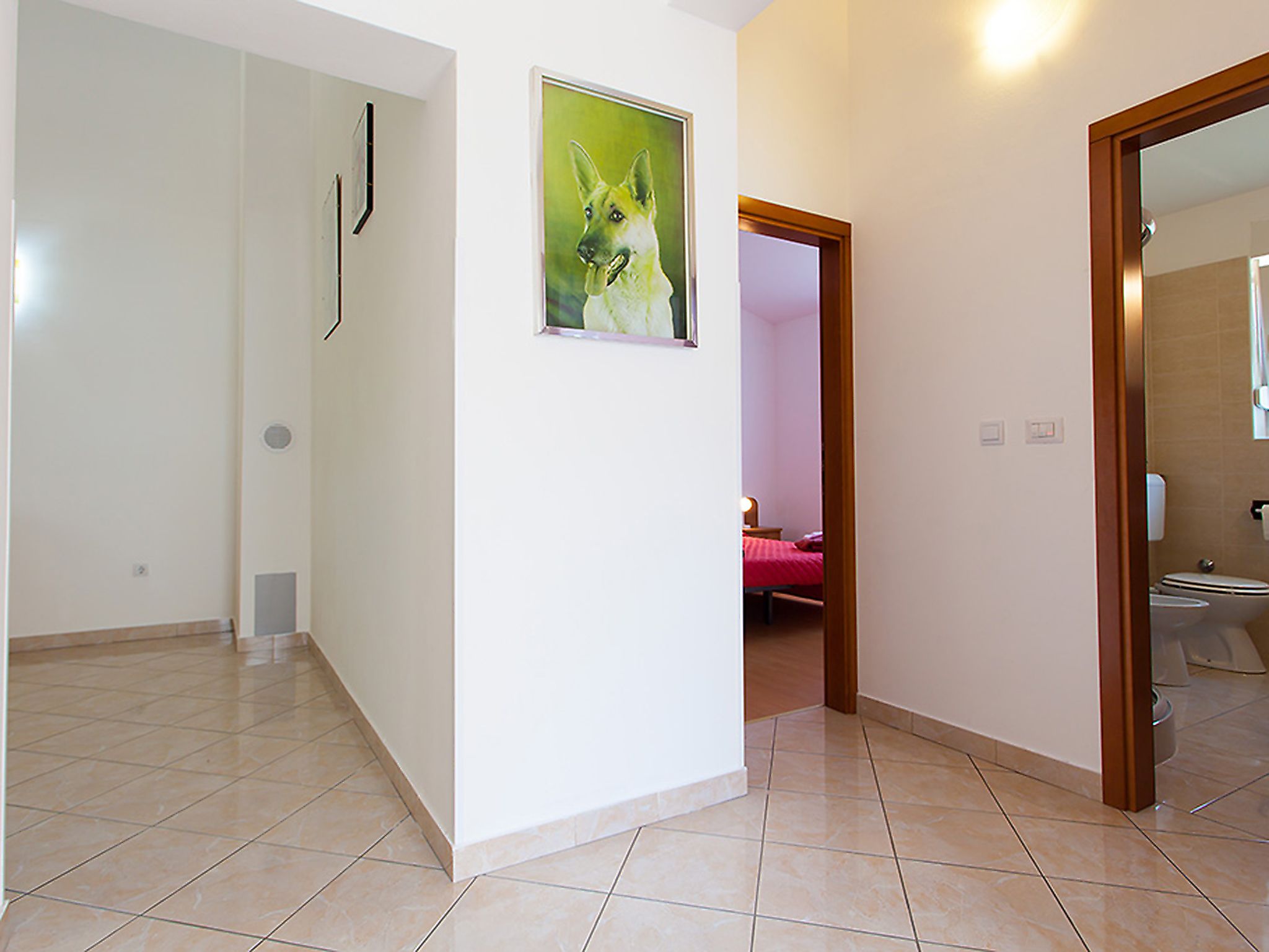 A hallway with a tiled floor, a painting of a dog, and a bathroom visible through an open door.