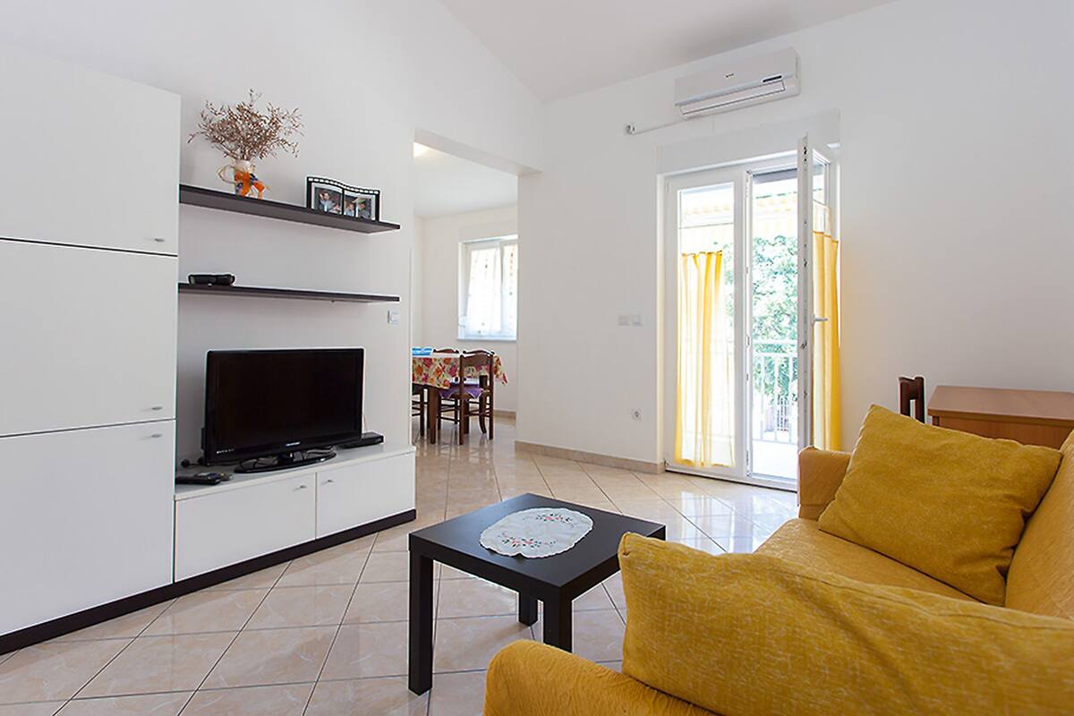 Uma sala de estar moderna com televisão de tela plana, uma área de jantar com mesa e cadeiras, e uma porta de vidro deslizante que dá para uma varanda.