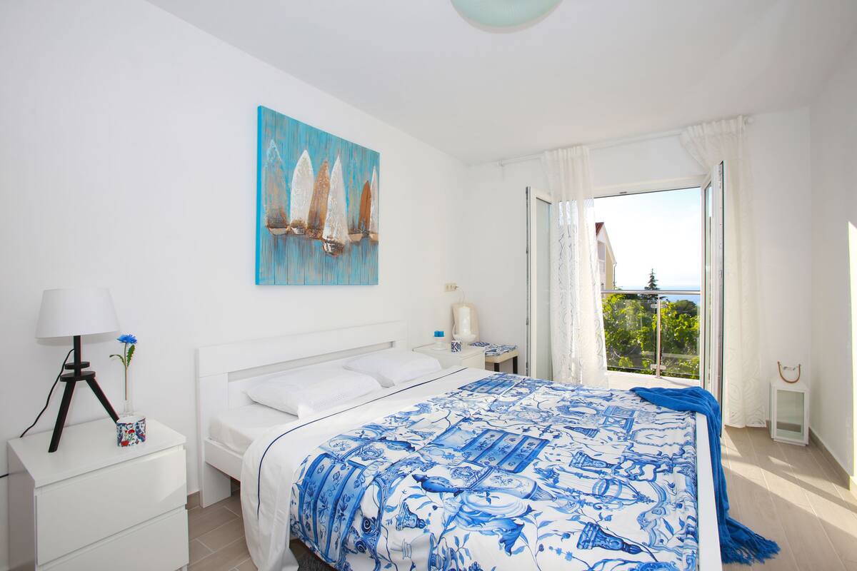Un dormitorio con una cama grande, una mesita de noche, una lámpara, un cuadro y vistas al exterior.