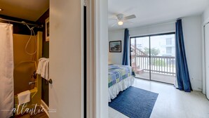 A bedroom with a bed, a window with a view, a ceiling fan, and a bathroom with a shower and a towel rack.