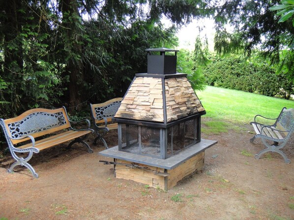 A rustic outdoor seating area with a stone-built wood stove, surrounded by benches and lush greenery.