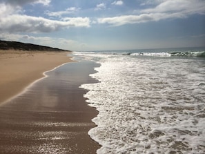 A beach with waves gently lapping onto the shore, a clear sky, and a distant shoreline. - Paradise on The Boulevard - 90 Mile Beach Holiday Home (Paradise Beach)