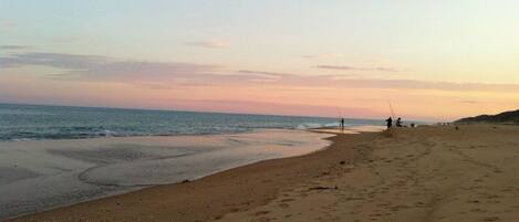 A beach with footprints leading to the water, a person fishing, and a distant shoreline.