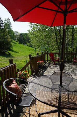 A covered outdoor seating area with a table and chairs, surrounded by a wooden deck and a red umbrella. - Best location in the Valley! Walk to weddings, concerts and mountain activities (Ellicottville)
