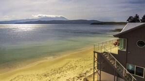 Une maison de plage dotée d’un balcon donnant sur l’océan, d’un escalier menant à l’étage supérieur et offrant une vue sur les montagnes lointaines.