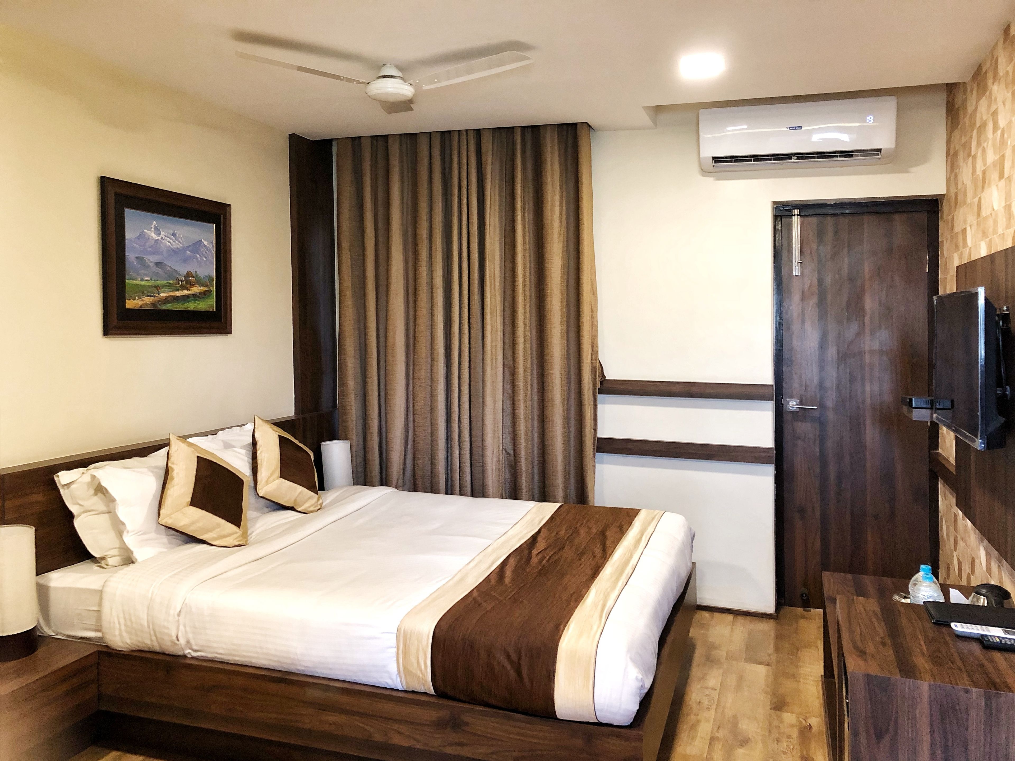 Habitación de hotel con cama, ventilador de techo, aire acondicionado, televisor montado en la pared y escritorio de madera con control remoto.