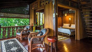 Une pièce en bois avec un lit, un ventilateur, des meubles en osier et une vue sur la verdure environnante.