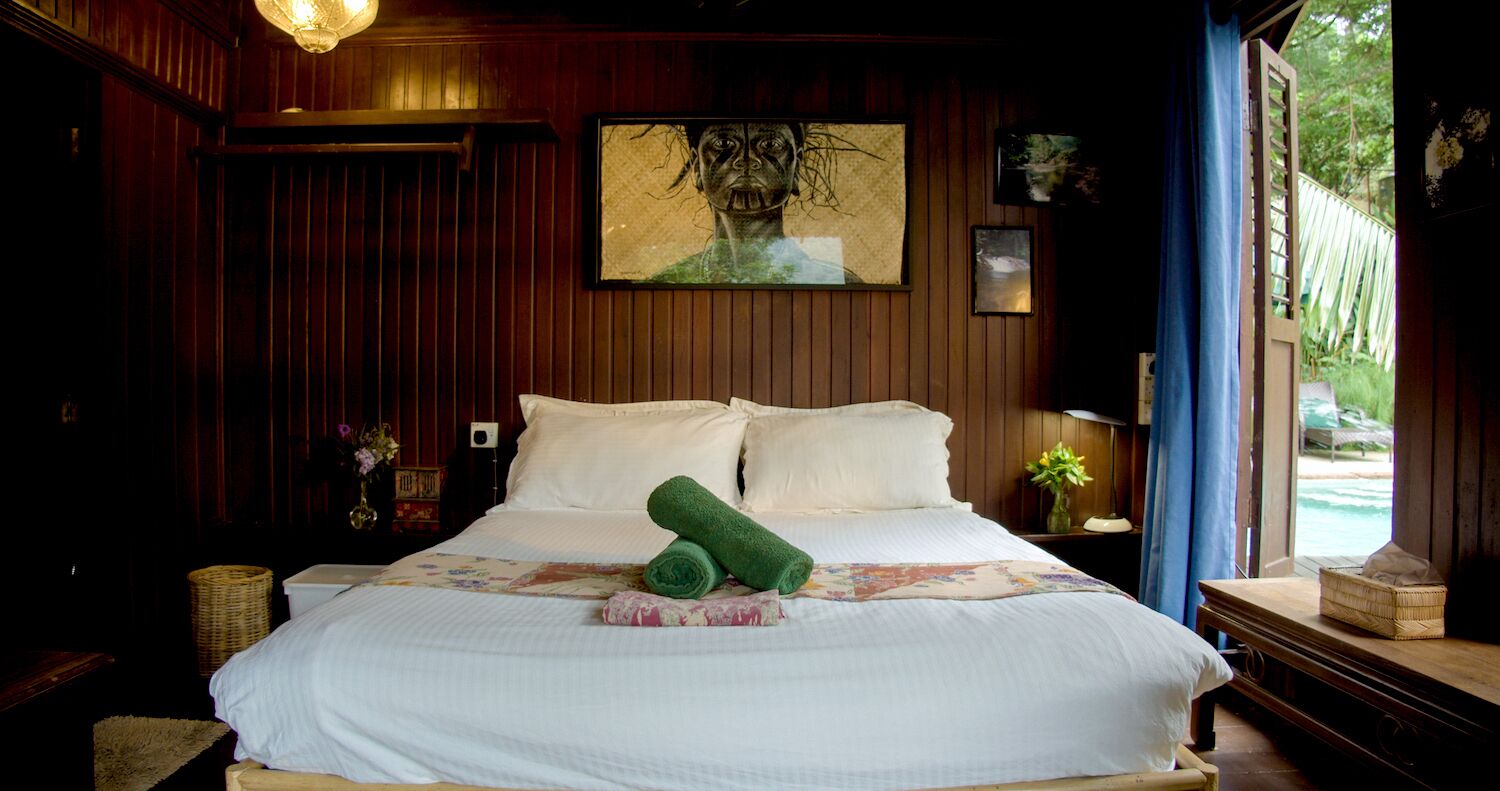 A wooden-paneled room with a bed, a framed artwork, a nightstand, a lamp, and a window with a view of greenery.