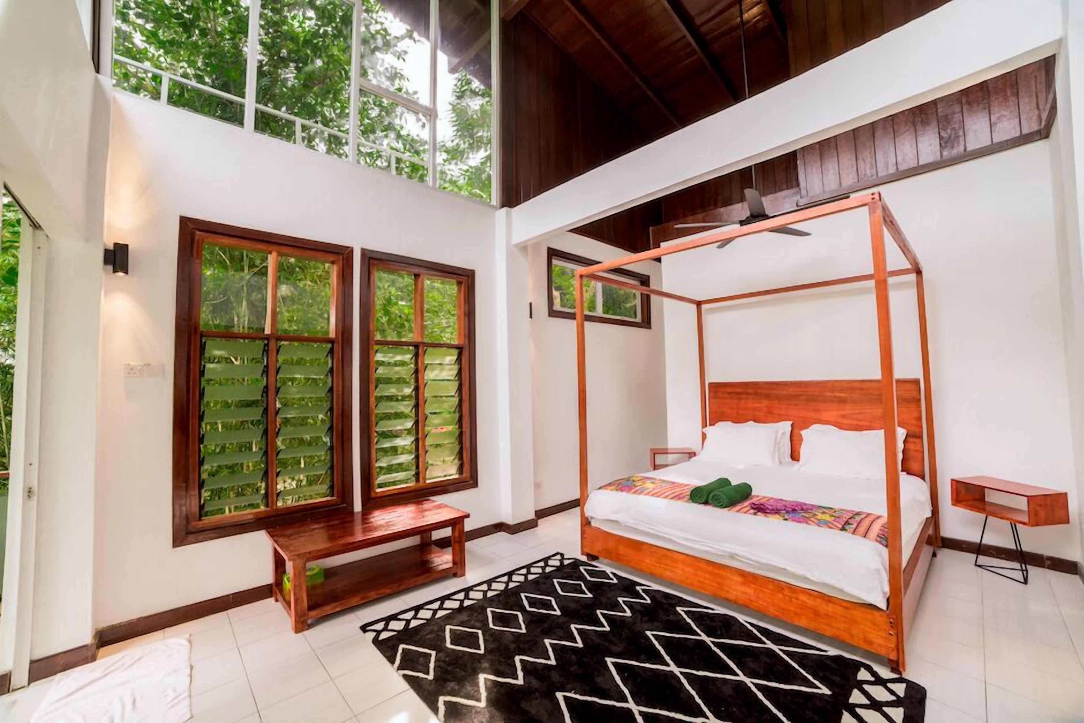 A bedroom with a four-poster bed, large windows with green foliage, a wooden bench, and a black and white rug.