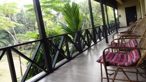 A covered outdoor area with a wooden floor, wicker chairs with striped cushions, and a view of lush greenery. - Rainbow Safari Resort (Sauraha)