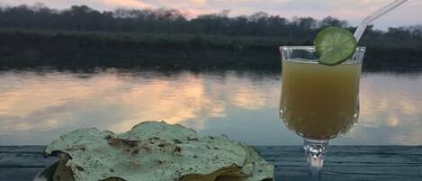 Un plato de pan plano y un vaso de bebida con una rodaja de lima sobre una mesa de madera junto a un lago al atardecer.