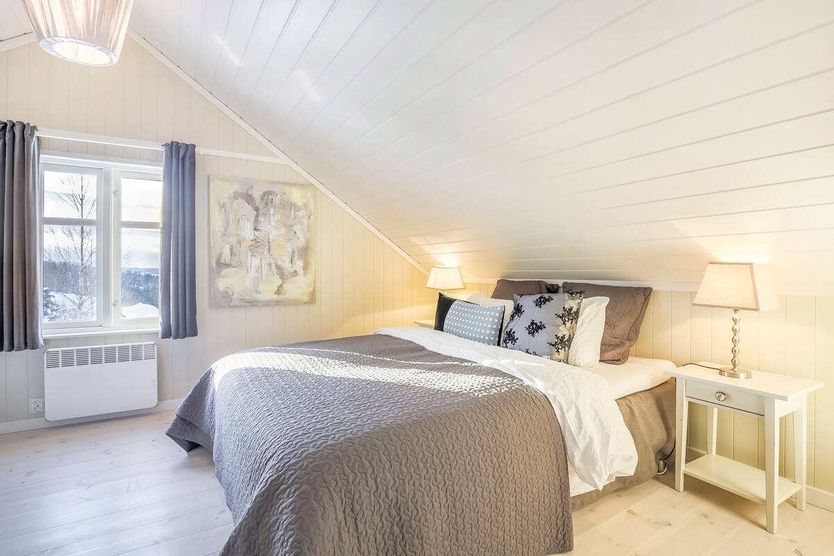 A bedroom with a sloped ceiling, a bed with a grey quilt, two bedside lamps, a white nightstand, and a window with curtains.
