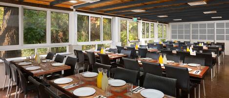 A spacious restaurant with multiple dining tables, black chairs, and large windows offering a view of greenery.