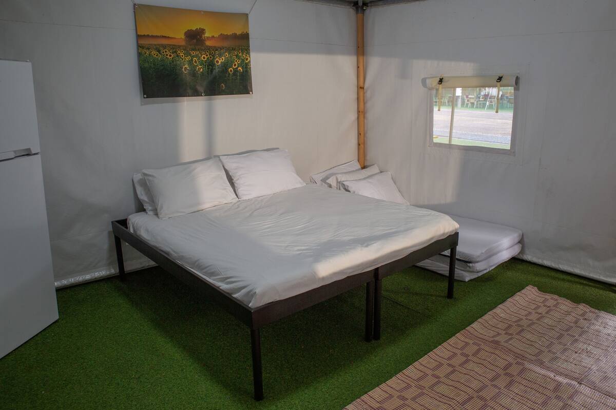 Ein Einzelbett mit weißer Bettwäsche in einem zeltofenartigen Raum mit grünem Boden und einem Gemälde einer Feldlandschaft im Abendrot.