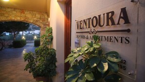 Letrero de 'VENTOURA ROOMS & APARTMENTS' con un arco de piedra y plantas en macetas al frente.
