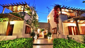 Un patio bien iluminado con un camino de piedra que lleva a un arco central, rodeado de exuberante vegetación y edificios blancos con detalles de madera.