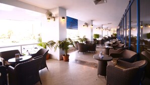 A spacious indoor seating area with wicker furniture and potted plants, large windows, and a blue awning. - Qasr Al Sahab (Khamis Mushait)