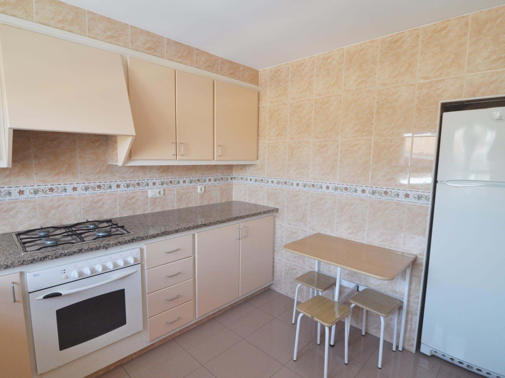 Eine Küche mit Gasherd, Backofen, weißen Schränken, einem Kühlschrank und einem kleinen Tisch mit zwei Stühlen.