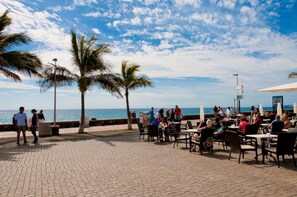 An outdoor café with tables and chairs, palm trees, and a view of the sea. - Meloneras Bahia 7 by VillaGranCanaria (Maspalomas)