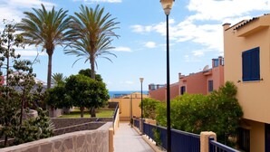 A pathway lined with palm trees and a stone wall, leading to a building with a blue railing and beige walls. - Meloneras Bahia 7 by VillaGranCanaria (Maspalomas)