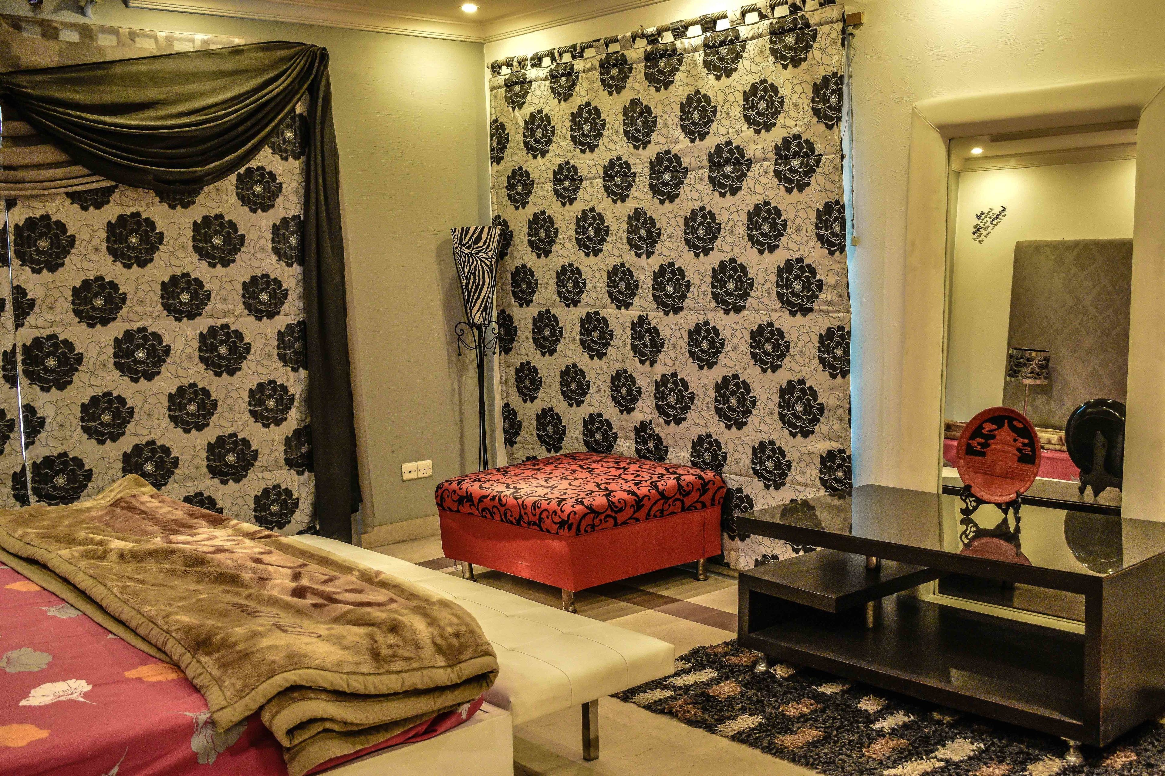 A bedroom with a bed, a red ottoman, a bedside table, and a mirror.