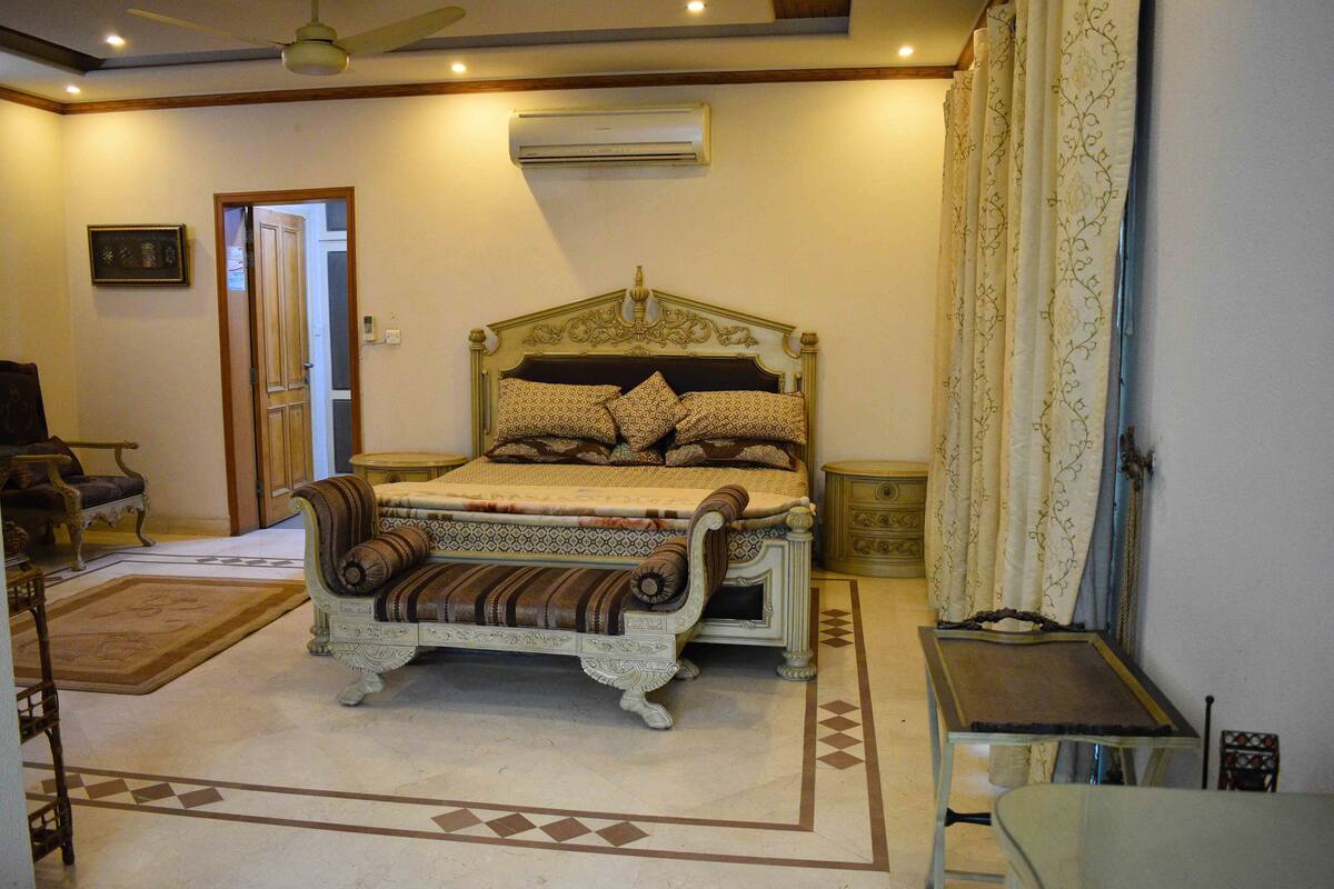 A bedroom with a large bed, a chair, a small table, and an air conditioning unit.