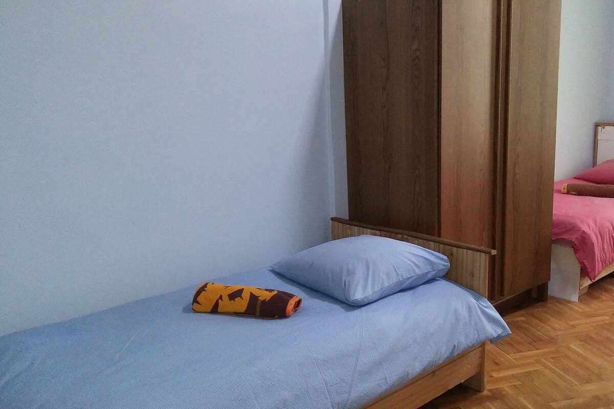 A small, single-bed room with a wooden wardrobe, a blue mattress, and a pink bedspread.