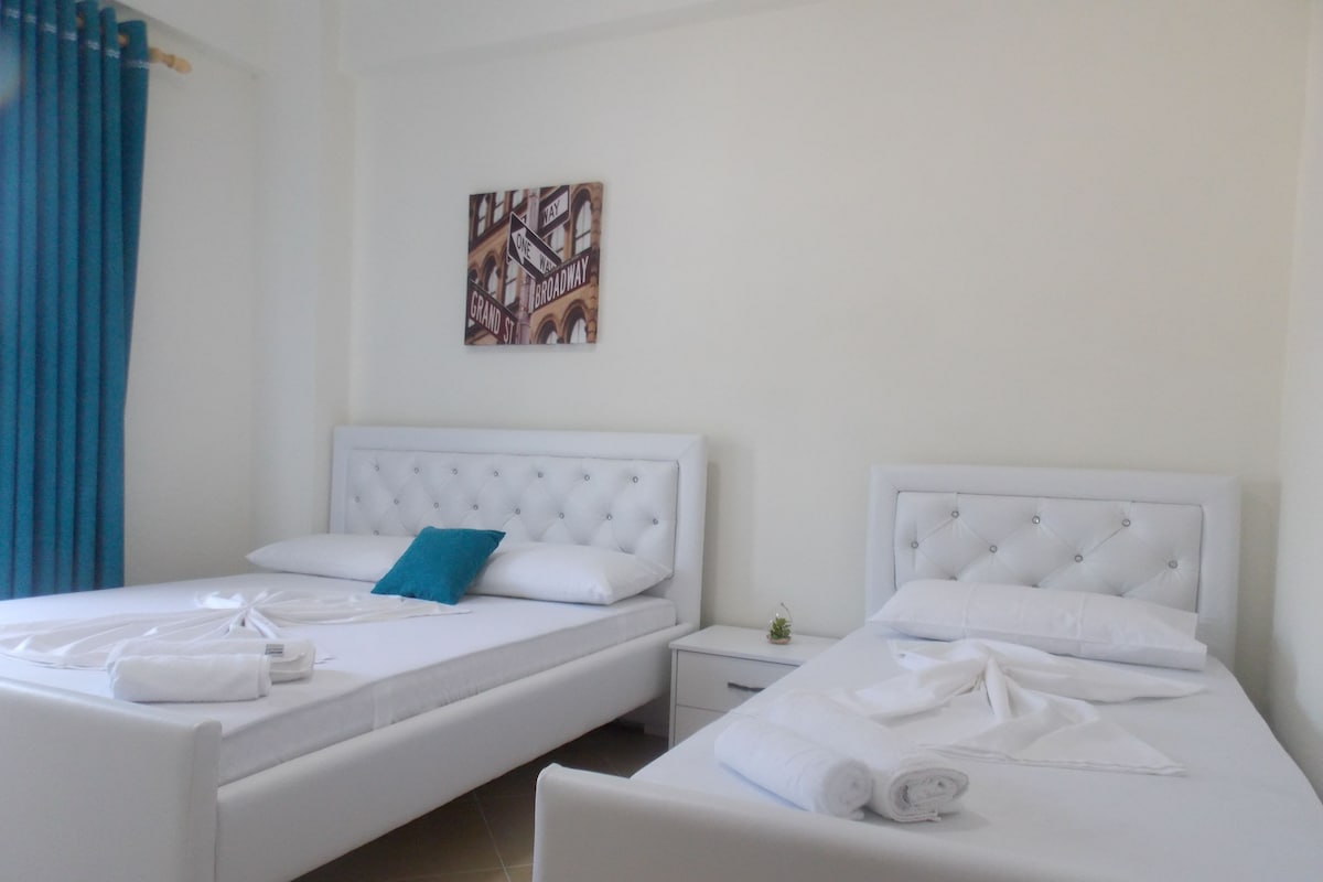 Una habitación de hotel con dos camas, cada una con ropa blanca y un cabecero capitonado. Se observa una mesita de noche con una lámpara y un adorno a un lado.