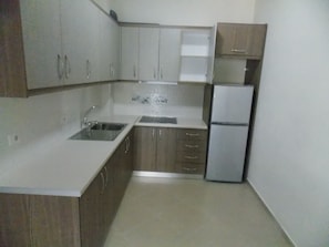 A compact kitchen with white countertops, wooden cabinets, a stainless steel sink, and a built-in refrigerator. - Doka Luxury Apartments (Sarandë)