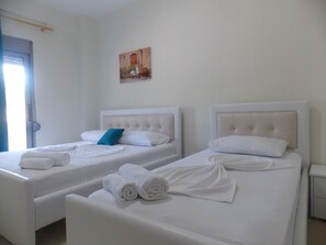 Two twin beds with white linens and rolled towels, a painting on the wall, and a door with a window. - Doka Luxury Apartments (Sarandë)