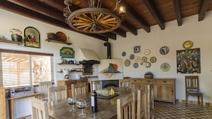 Un comedor rústico con mesa de madera, sillas y una araña. Se observan varios utensilios de cocina y platos decorativos en las paredes.