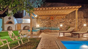 A covered outdoor seating area with stone walls, a pool, and lounge chairs.