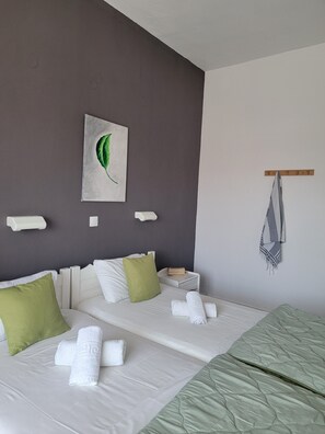 Camera d'albergo con due letti, un cuscino verde, un cuscino bianco, un quadro raffigurante una foglia, un portasciugamani e un comodino con dei libri.