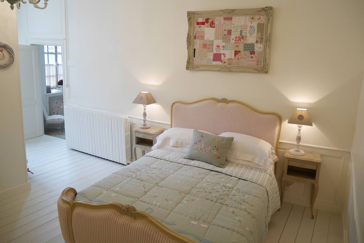 A bedroom with a bed, two bedside tables, a nightstand, a chandelier, a wall hanging, and a window.