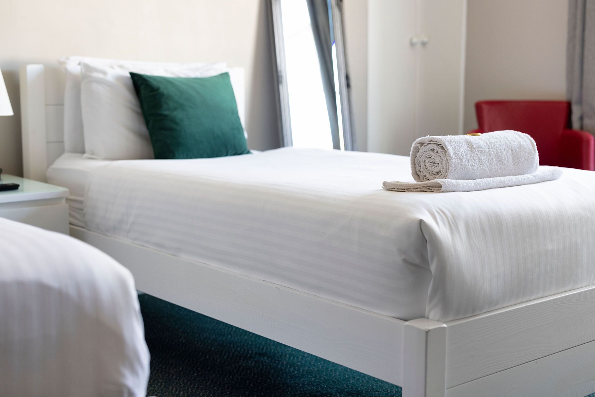 A hotel room with a neatly made bed, a red chair, and a white nightstand with a lamp.