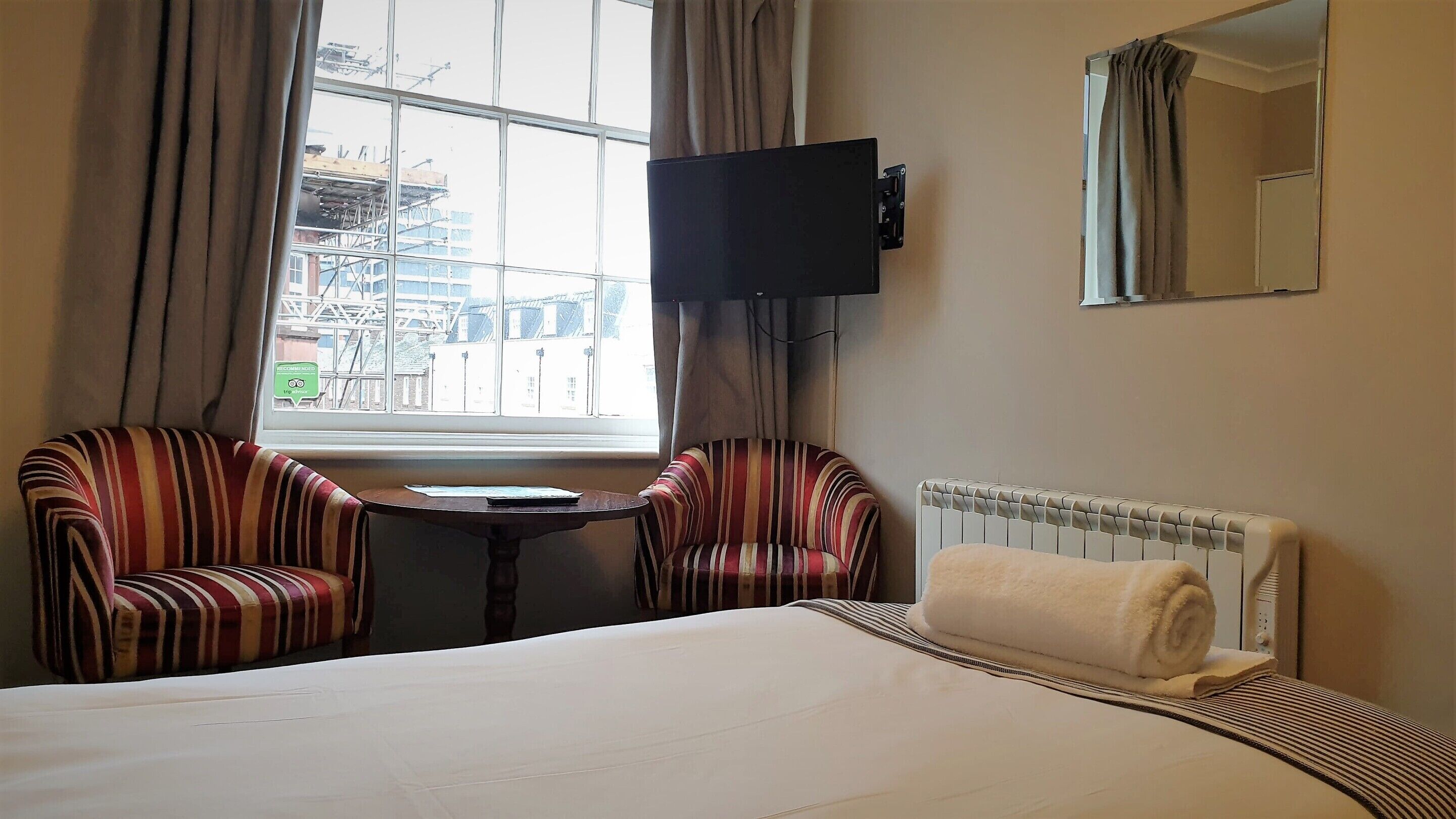 Een hotelkamer met een bed, twee gestreepte fauteuils, een tafeltje, een aan de muur bevestigde televisie en een raam met gordijnen.