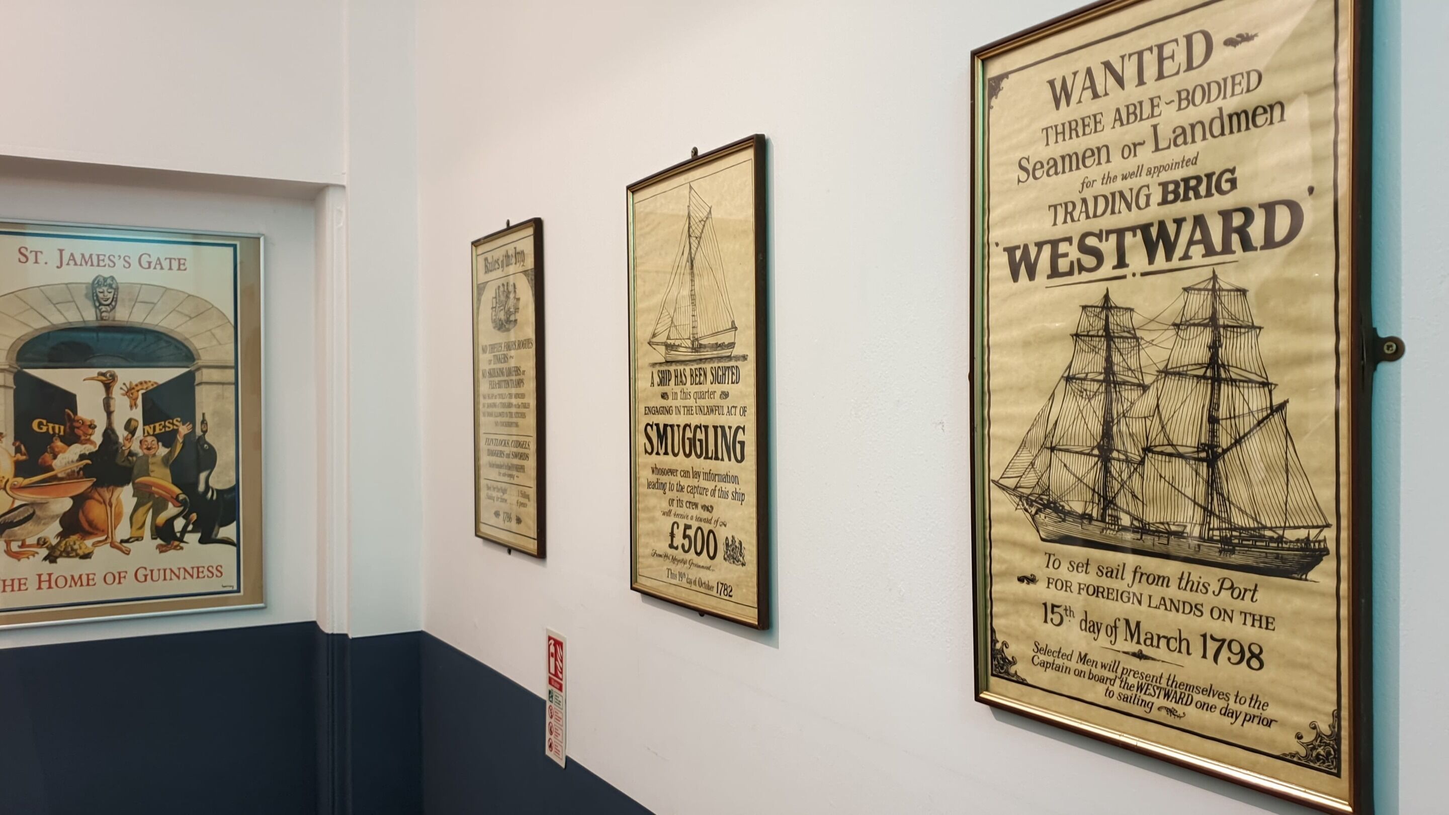 Una serie di manifesti storici su una parete, tra cui uno raffigurante una nave chiamata 'WESTWARD' e un altro che fa riferimento a 'St. James's Gate'.
