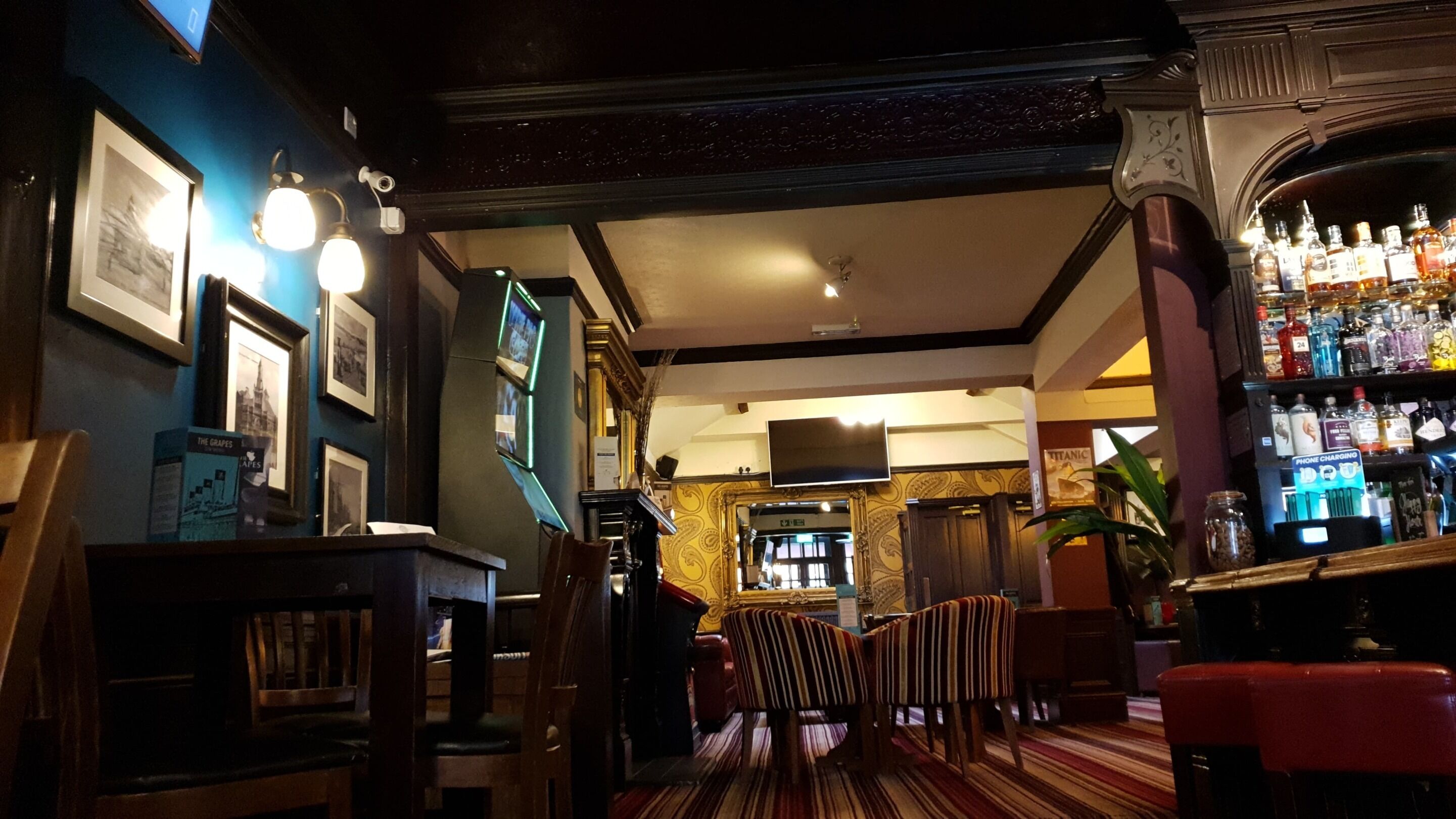 A bar area with a wooden bar counter, multiple framed pictures on the wall, a seating area with striped chairs, and a bar stocked with various bottles.