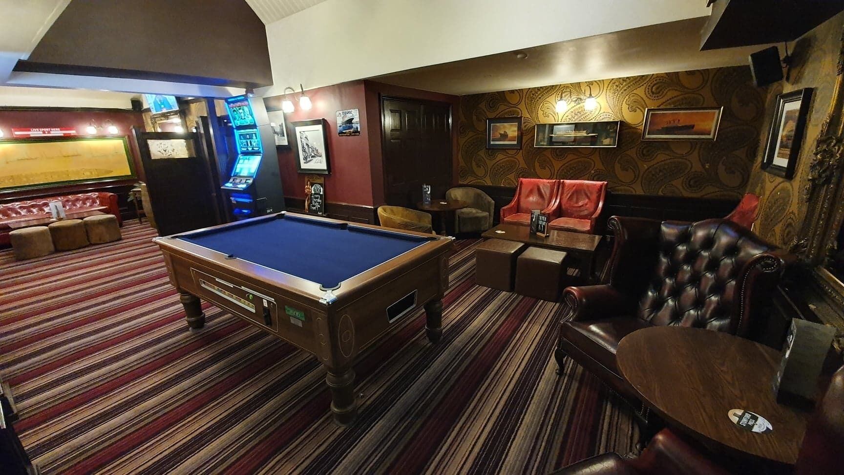 A pool table in a room with striped carpet, a bar area with neon lights, and leather seating.