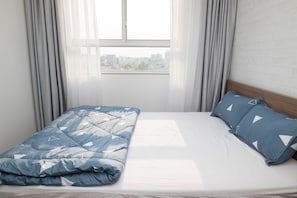 Un lit avec une couette et des oreillers bleus, une fenêtre avec des rideaux et une vue sur la ville.