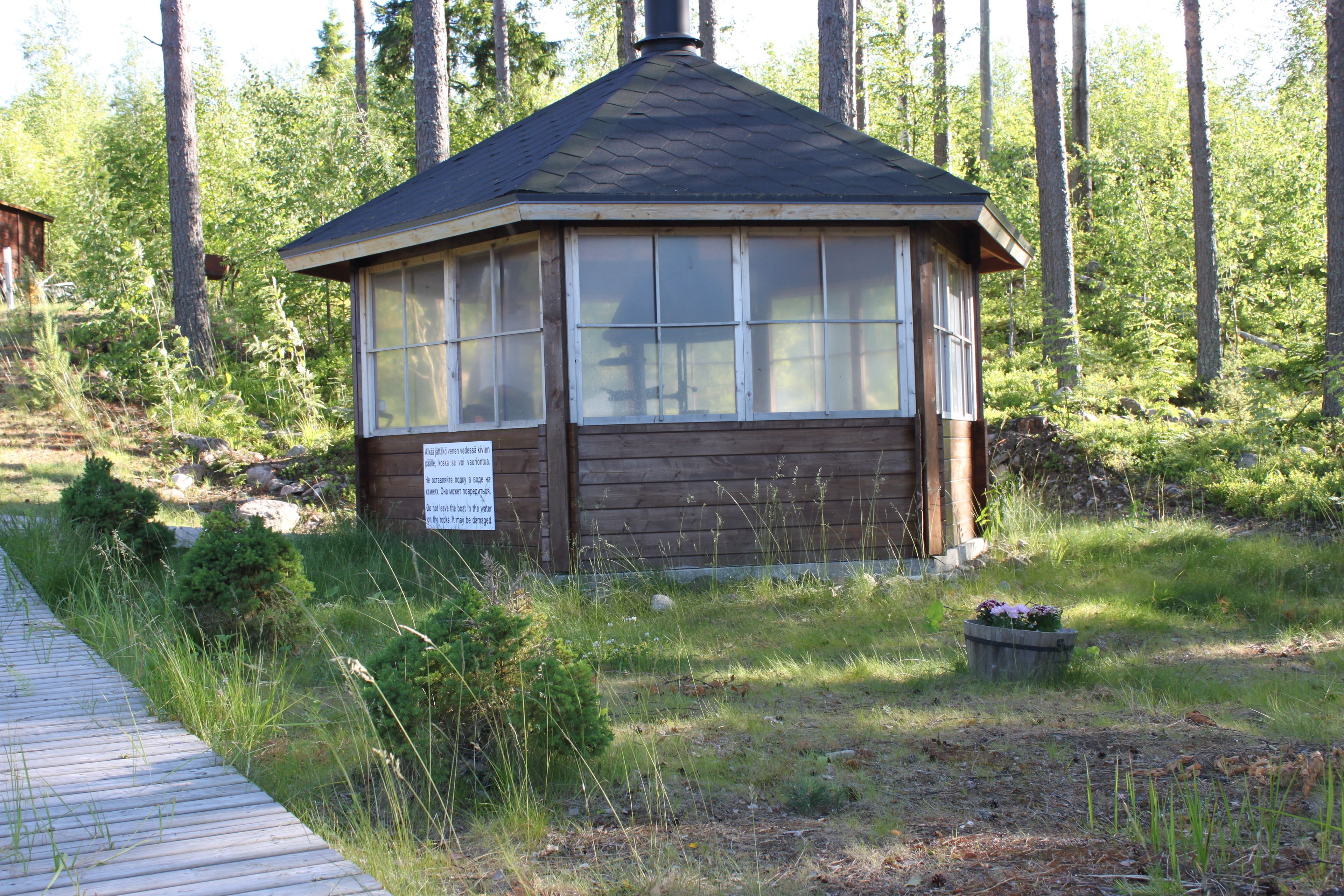 Puinen mökki, jossa on harjakatto ja suuret ikkunat, ja sen ympärillä on metsää.