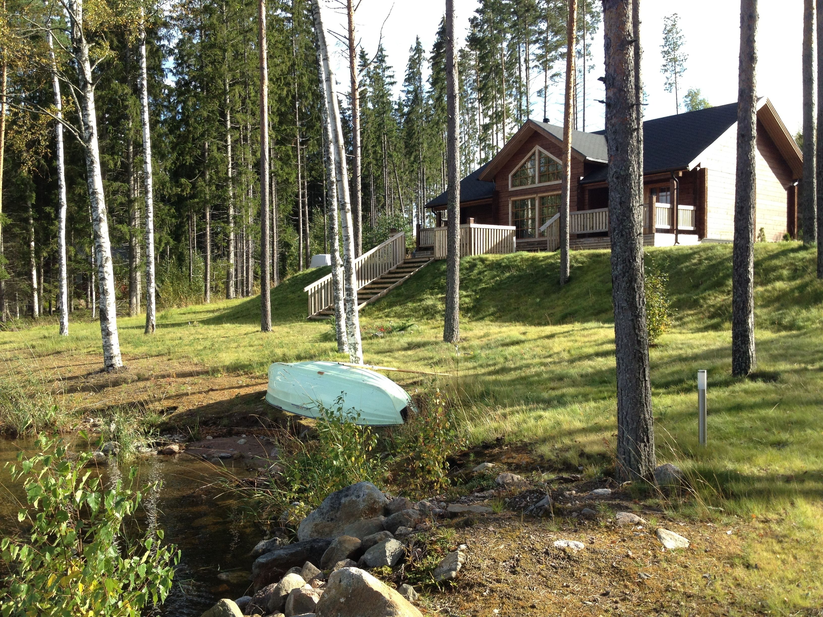 Une cabane dotée d’une terrasse en bois et d’un escalier menant à un porche, le tout entouré de grands arbres et traversé par un petit ruisseau.