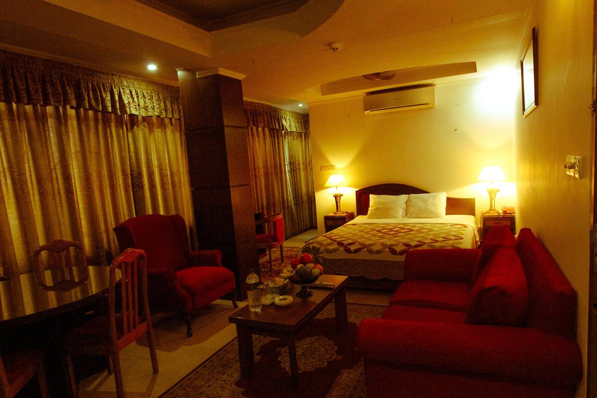 A hotel room with a bed, two red armchairs, a wooden coffee table, and a desk with a chair.