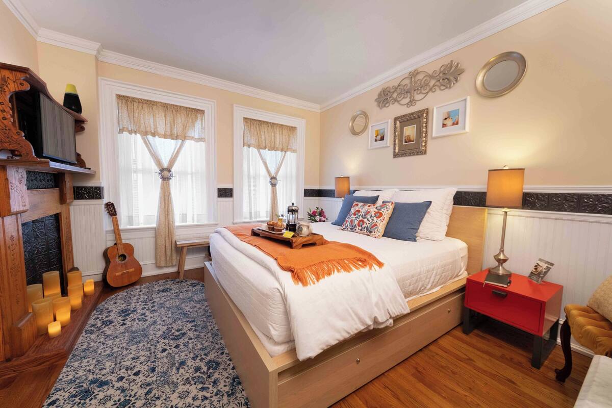 A bedroom with a bed, bedside tables, a lamp, a guitar, and a fireplace.