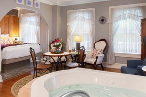 A room with a bed, a sitting area with a table and chairs, a bathtub, and large windows with curtains. - Main Street Inn (Parkville)