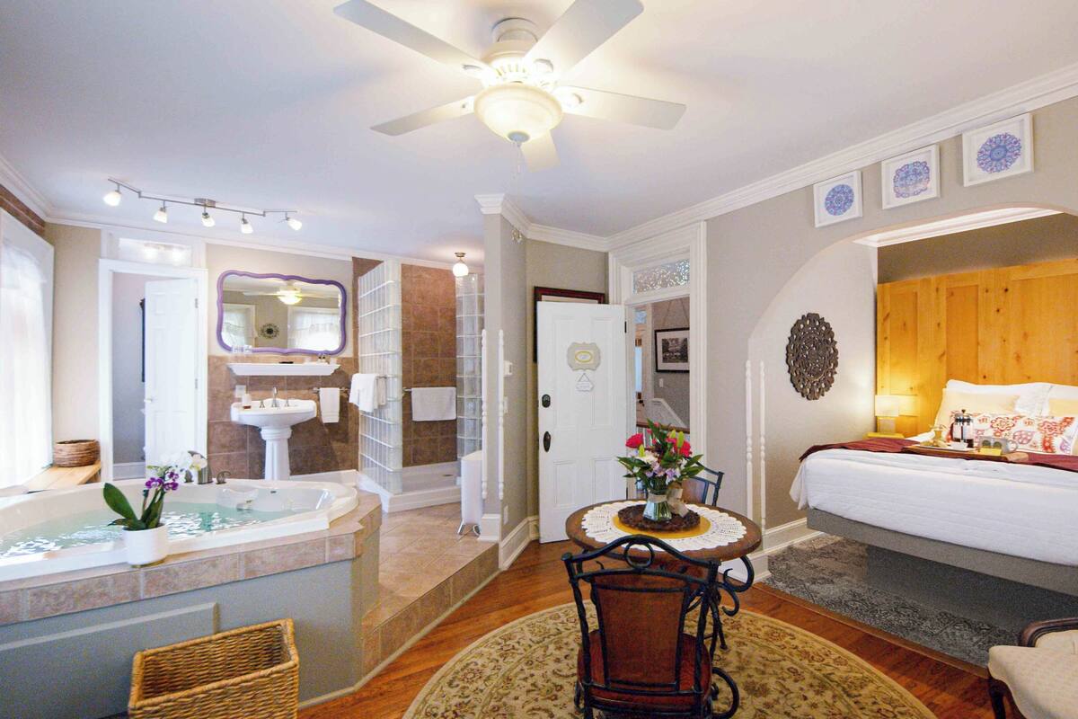 A hotel room with a large bed, a sitting area with a table and chairs, a bathroom with a bathtub, and a ceiling fan.
