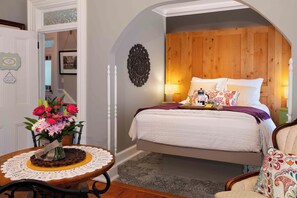 A bedroom with a bed, bedside lamps, a wooden headboard, a small table with a vase of flowers, and a chair. - Main Street Inn (Parkville)