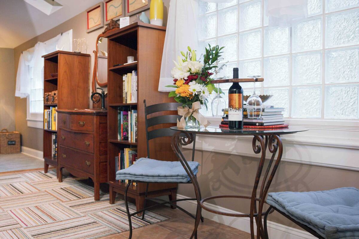 A bedroom with a bed, a wooden dresser, a chair, a small table with a bottle, and a window with a view.