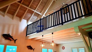 Una habitación con techos altos, vigas de madera, un balcón con barandillas negras y dos lámparas colgantes.