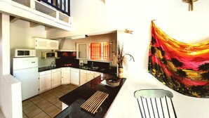Una cocina compacta con gabinetes blancos, un microondas y un refrigerador. Hay una zona de comedor con una encimera negra y una silla. Un cuadro colorido decora la pared.